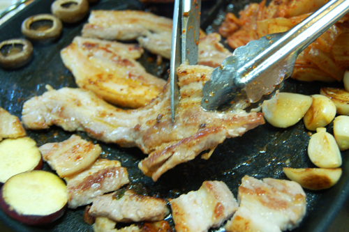 強火で熱した鉄板に（1）と白菜キムチをのせ、肉がこんがり焼けてきたら裏返し、豚肉がカリカリになるまで焼く。マッシュルームは傘を下にしてのせ、片面だけ焼く。白菜キムチ、にんにく、さつまいもは、焦げないよう気をつけながら両面焼く。焼き上がったら、豚肉と白菜キムチをハサミで一口大に切る。