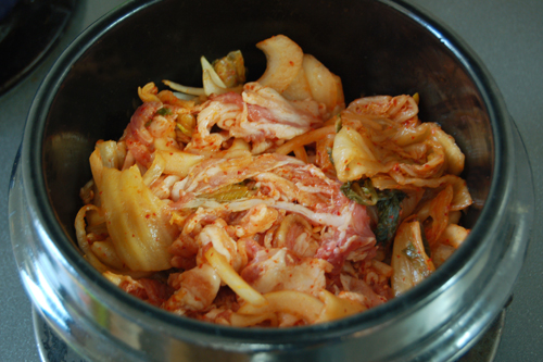 トゥッペギ（または土鍋）に、キムチ、豚肉、玉ねぎを入れて混ぜ合わせ、10分ほどおいてから火にかけ、2分ほど炒める。粉唐辛子、酒、砂糖を加え、弱火でさらに1～2分炒める。